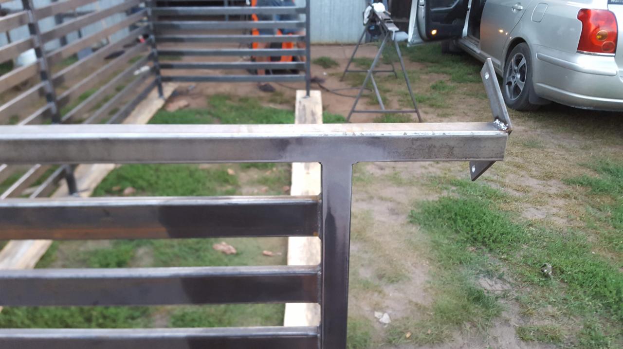 Spawana metalowa konstrukcja ogrodzenia w trakcie montażu z widocznymi elementami mocującymi, spawarką w tle i samochodem z otwartymi drzwiami.