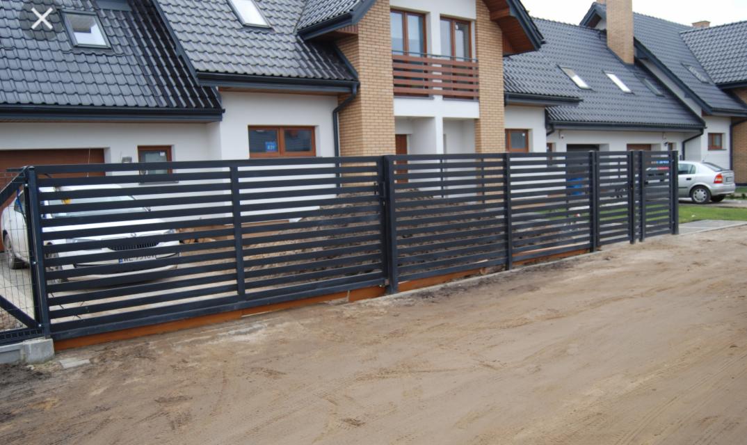 Nowoczesne, poziome ogrodzenie metalowe w kolorze grafitowym przed nowo wybudowanym domem jednorodzinnym z szarą dachówką, widoczne samochody za ogrodzeniem.