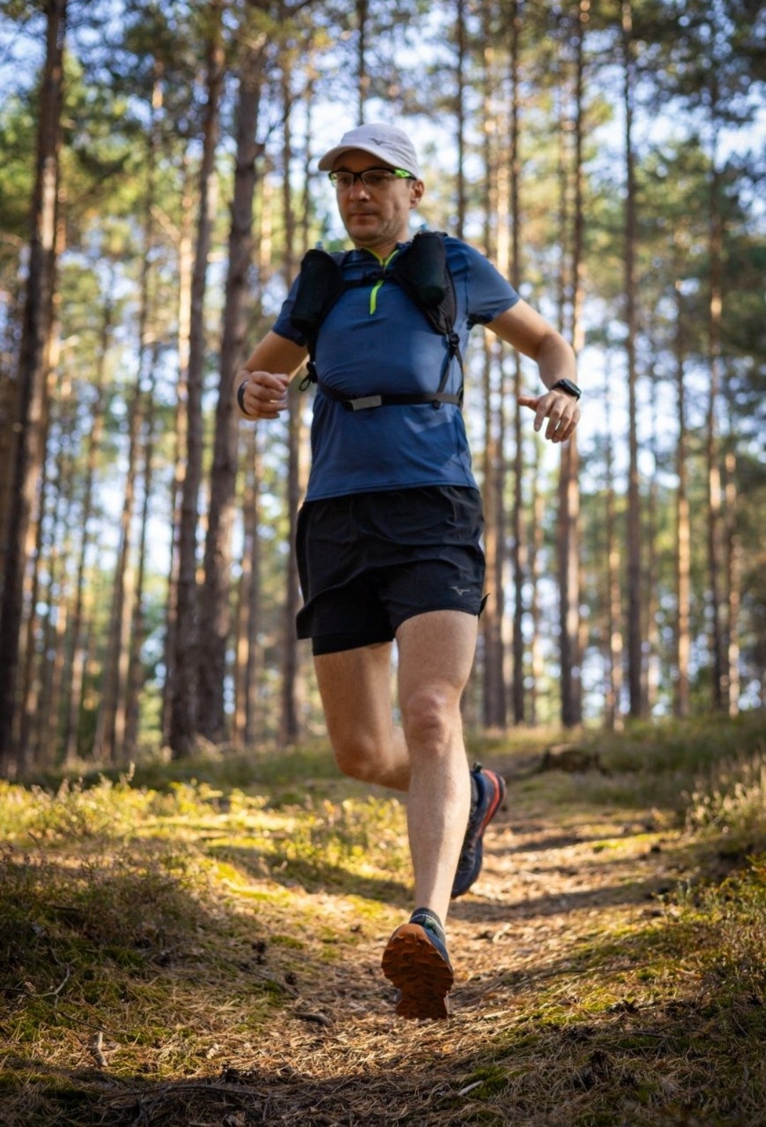 Mężczyzna w okularach i sportowym stroju biegnie ścieżką w lesie, widoczne drzewa i ściółka leśna.