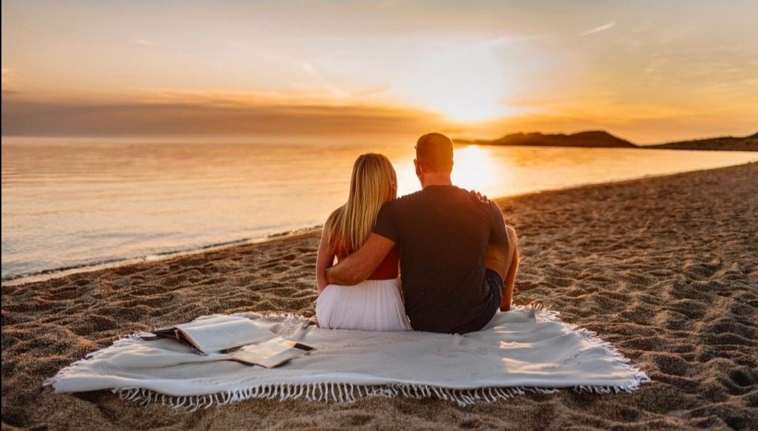 Para siedzi na kocu na plaży o zachodzie słońca, czytając książkę.