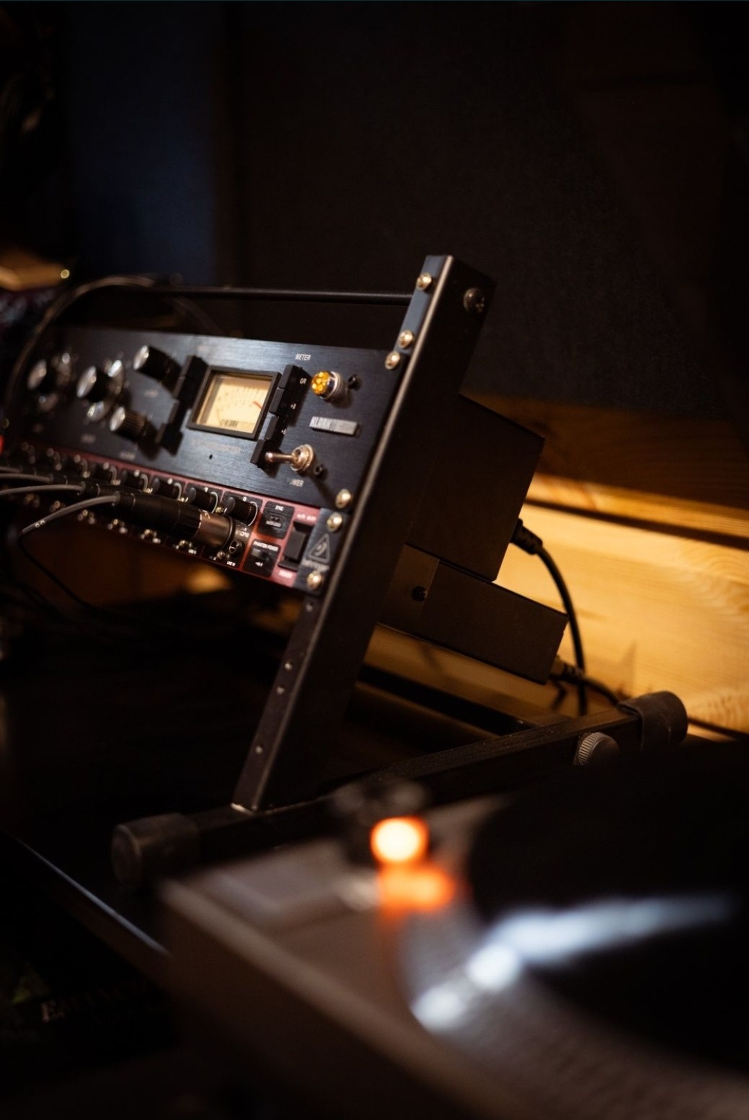 Czarny rack audio z analogowymi wskaźnikami i kablami, ustawiony na metalowym stojaku, z gramofonem widocznym w tle w przyciemnionym studio.