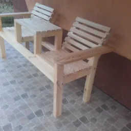 Drewniana ławka parkowa z dwoma siedziskami i małym stolikiem pośrodku, wykonana z jasnego drewna. Widoczne detale konstrukcyjne i naturalna faktura drewna.