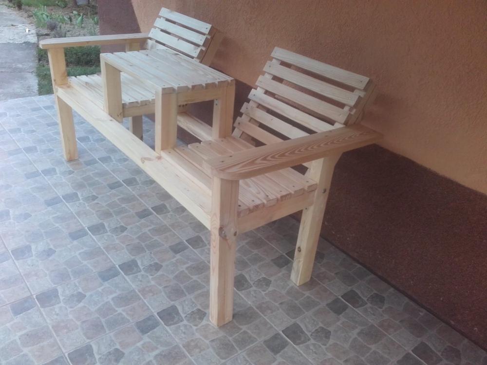 Drewniana ławka parkowa z dwoma siedziskami i małym stolikiem pośrodku, wykonana z jasnego drewna. Widoczne detale konstrukcyjne i naturalna faktura drewna.