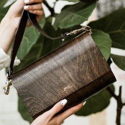 URBIK Photography - Marcin Łubianka - Kobieta prezentuje drewnianą torebkę marki Zisou na tle zielonych liści. Widoczne dłonie z białym manicure i fragment białej bluzki. Elegancki dodatek.