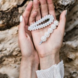 URBIK Photography - Marcin Łubianka - Dwie spinki do włosów z perłami w dłoniach kobiety na tle kory drzewa. Biały manicure i bluzka z falbankami dodają elegancji. Delikatne światło podkreśla detale.