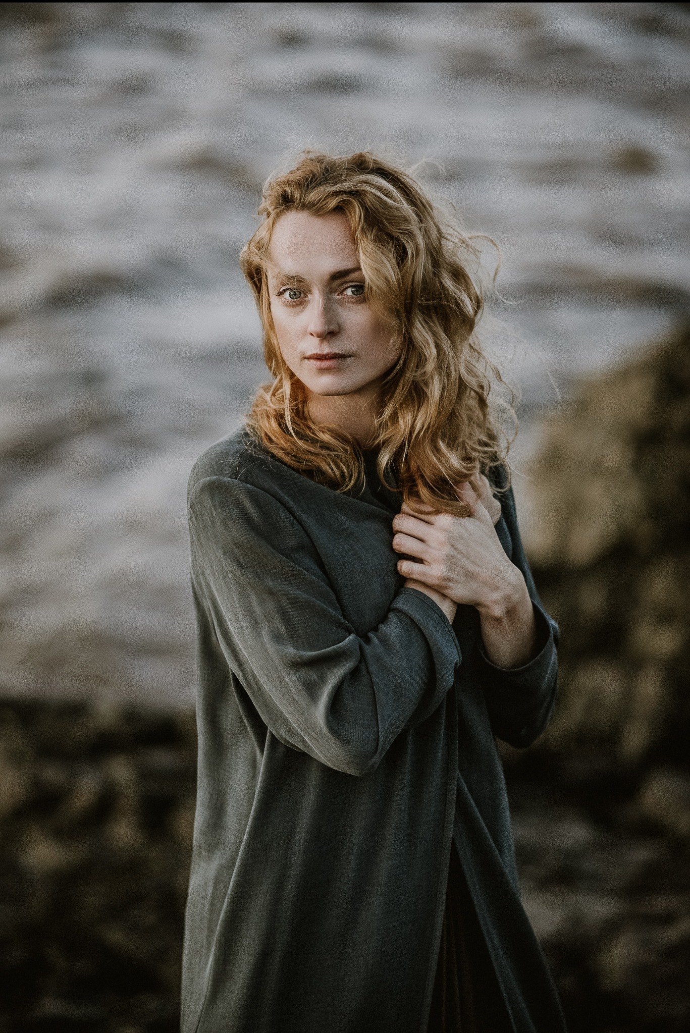 Portret kobiety o blond włosach w szaro-zielonej sukience na tle rozmytego krajobrazu. Delikatne światło podkreśla naturalność i subtelność ujęcia.