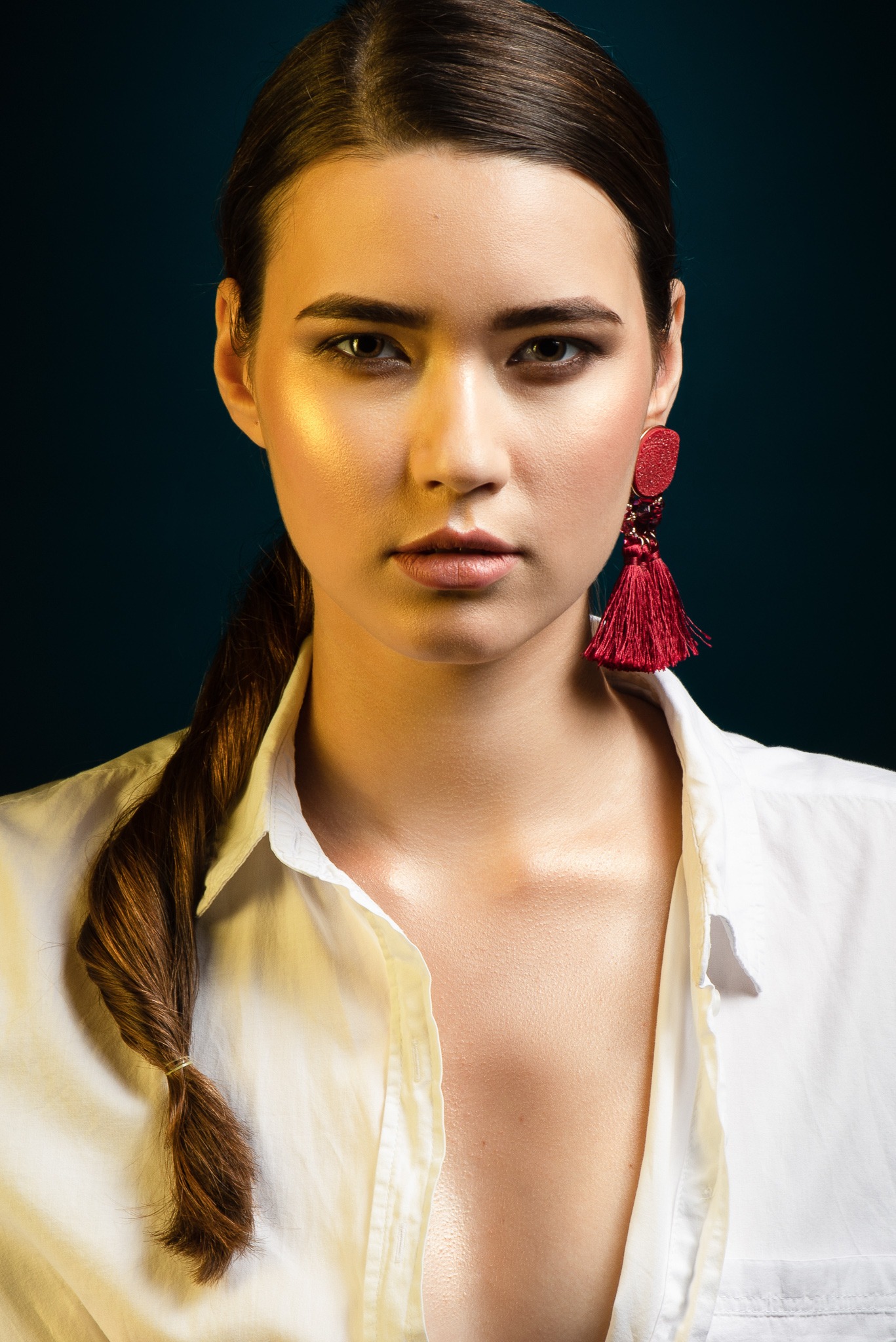Portret kobiety w białej koszuli z czerwonym kolczykiem na ciemnym tle. Uczesane włosy, profesjonalny makijaż i oświetlenie podkreślają urodę modelki.