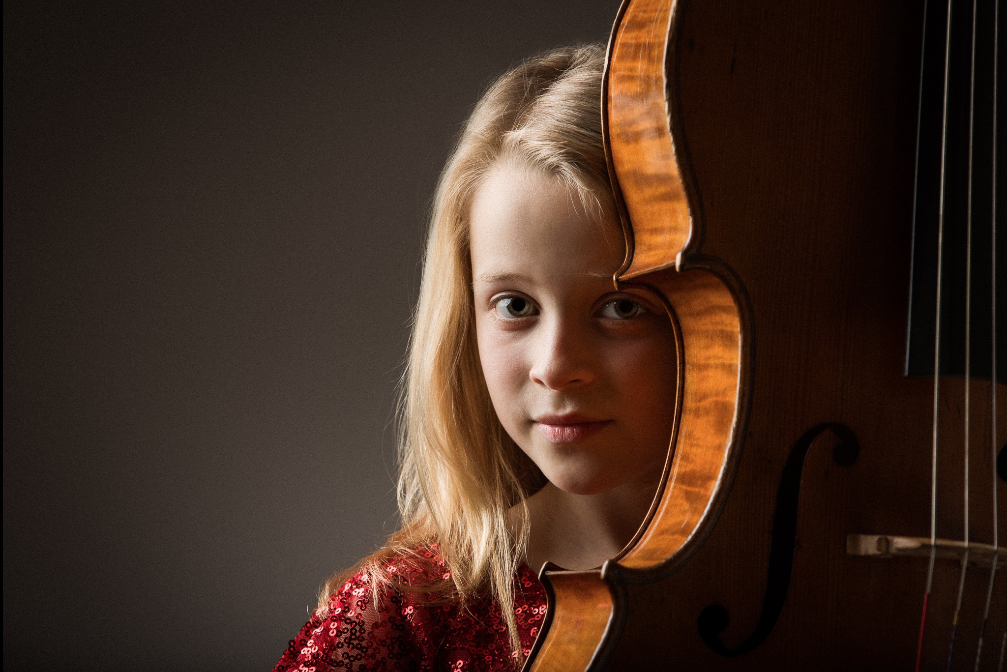 Portret dziewczynki z blond włosami, w czerwonej cekinowej sukience, pozującej z wiolonczelą na ciemnym tle. Ujęcie z bliska, skupione na twarzy i instrumencie.