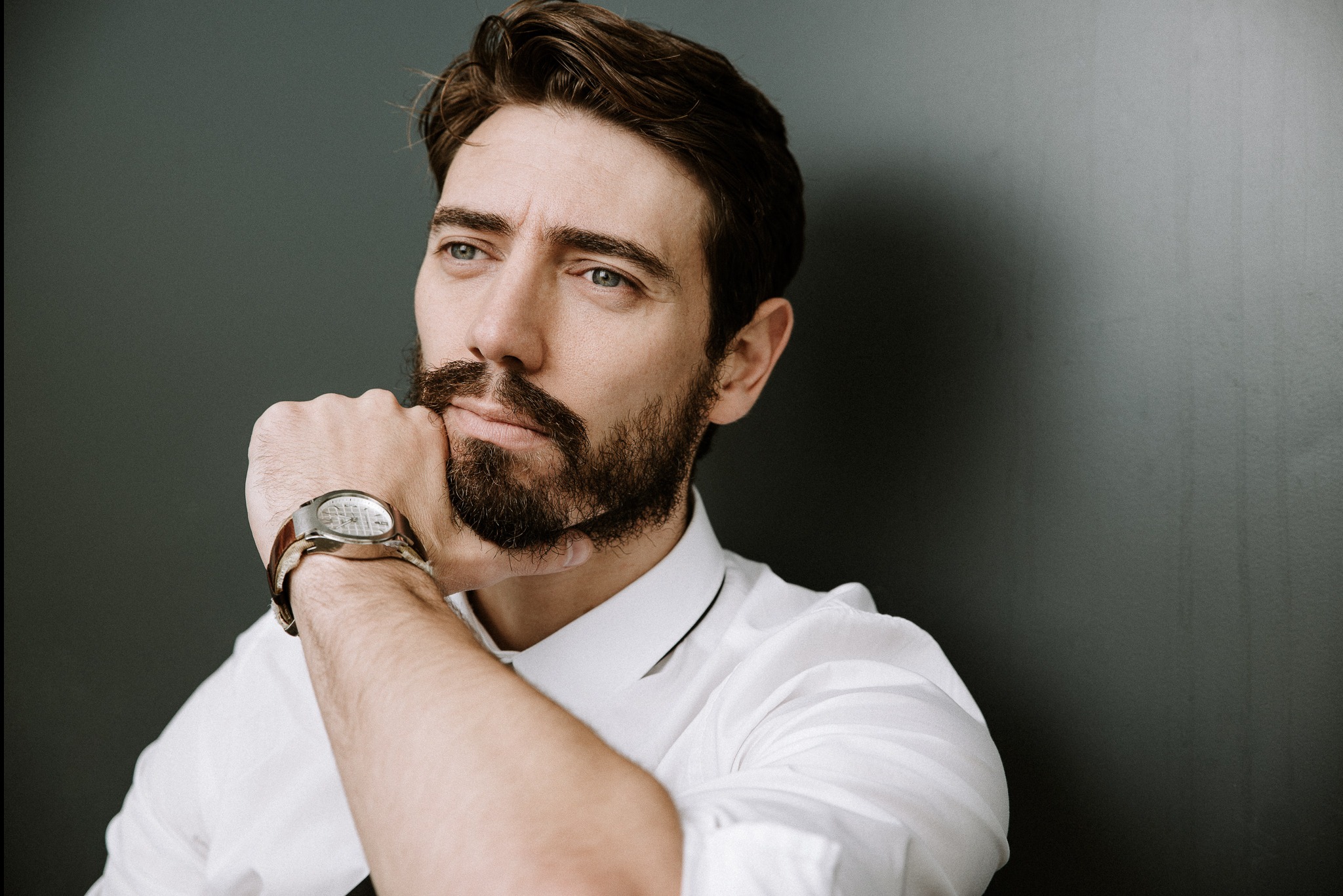 Portret mężczyzny z brodą w białej koszuli, z zegarkiem na ręku, opierającego podbródek na dłoni, zamyślony wzrok skierowany w lewo, na ciemnym tle.