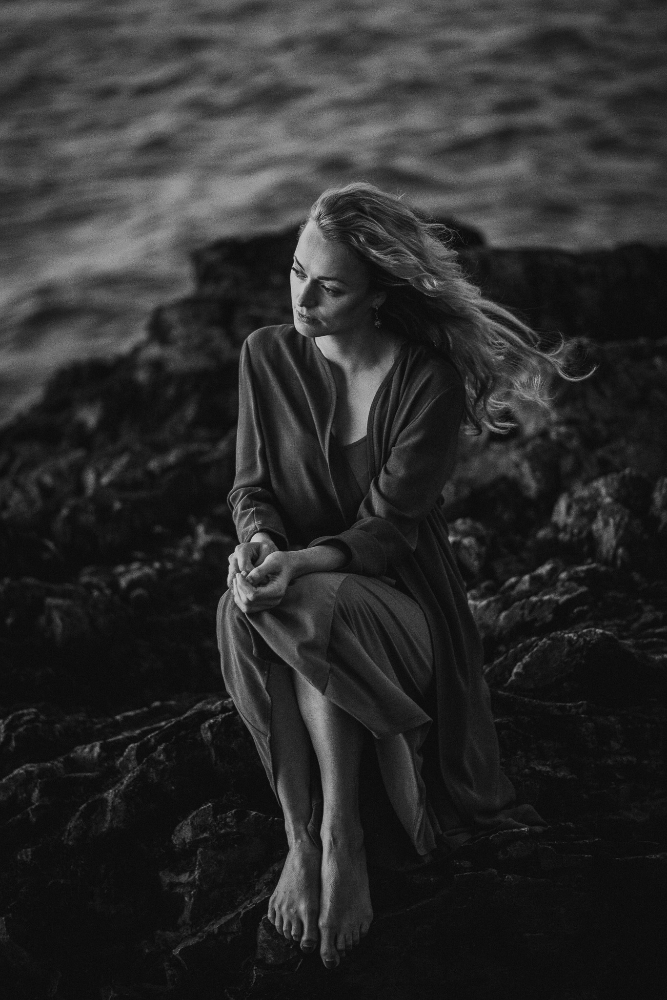 Czarno-białe zdjęcie kobiety siedzącej na skałach nad morzem, włosy rozwiane na wietrze, melancholijny wyraz twarzy, artystyczna fotografia portretowa.