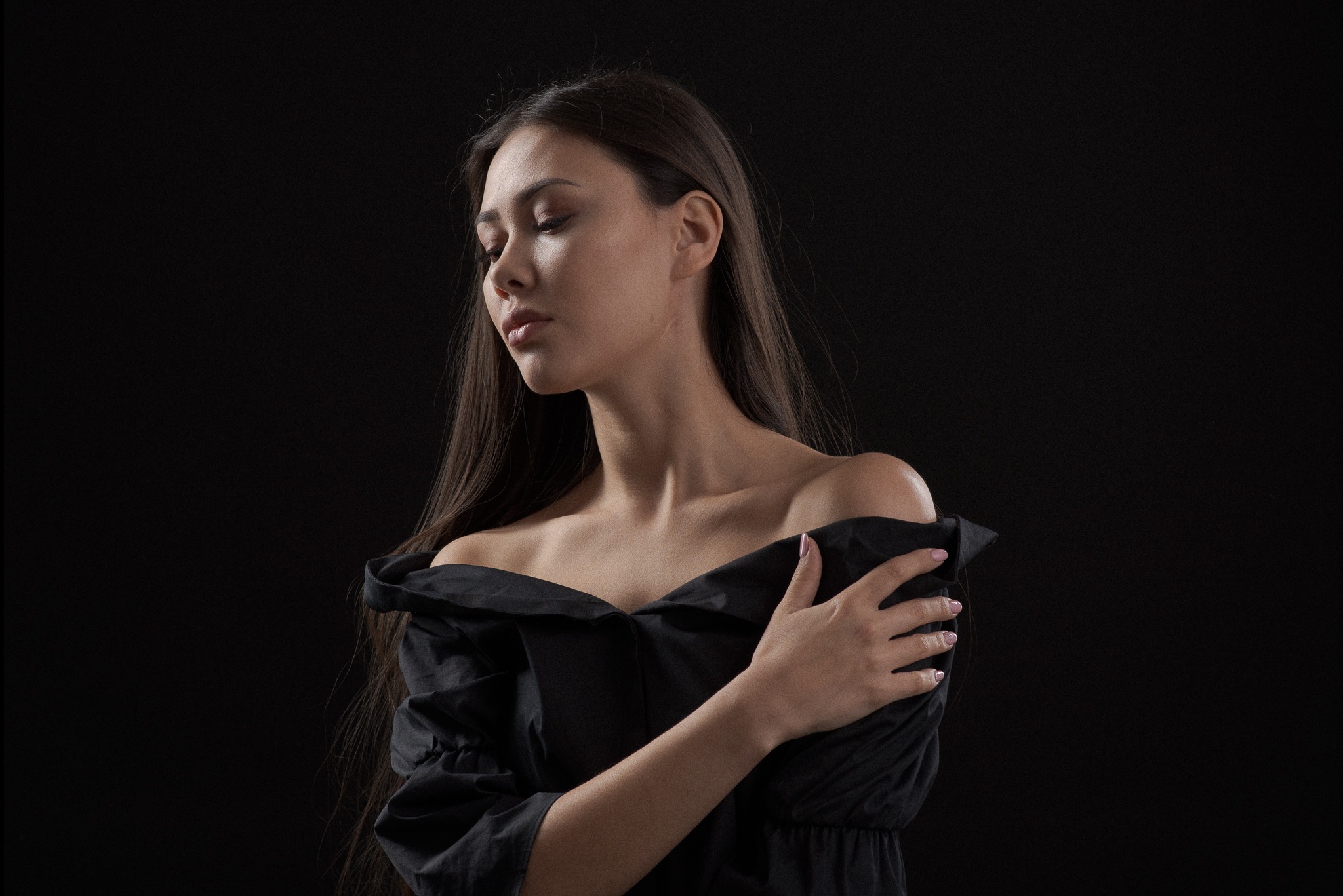 Portret kobiety w czarnej sukience z odkrytymi ramionami na ciemnym tle. Modelka patrzy w dół, z ręką skrzyżowaną na piersi. Subtelne oświetlenie podkreśla rysy twarzy.