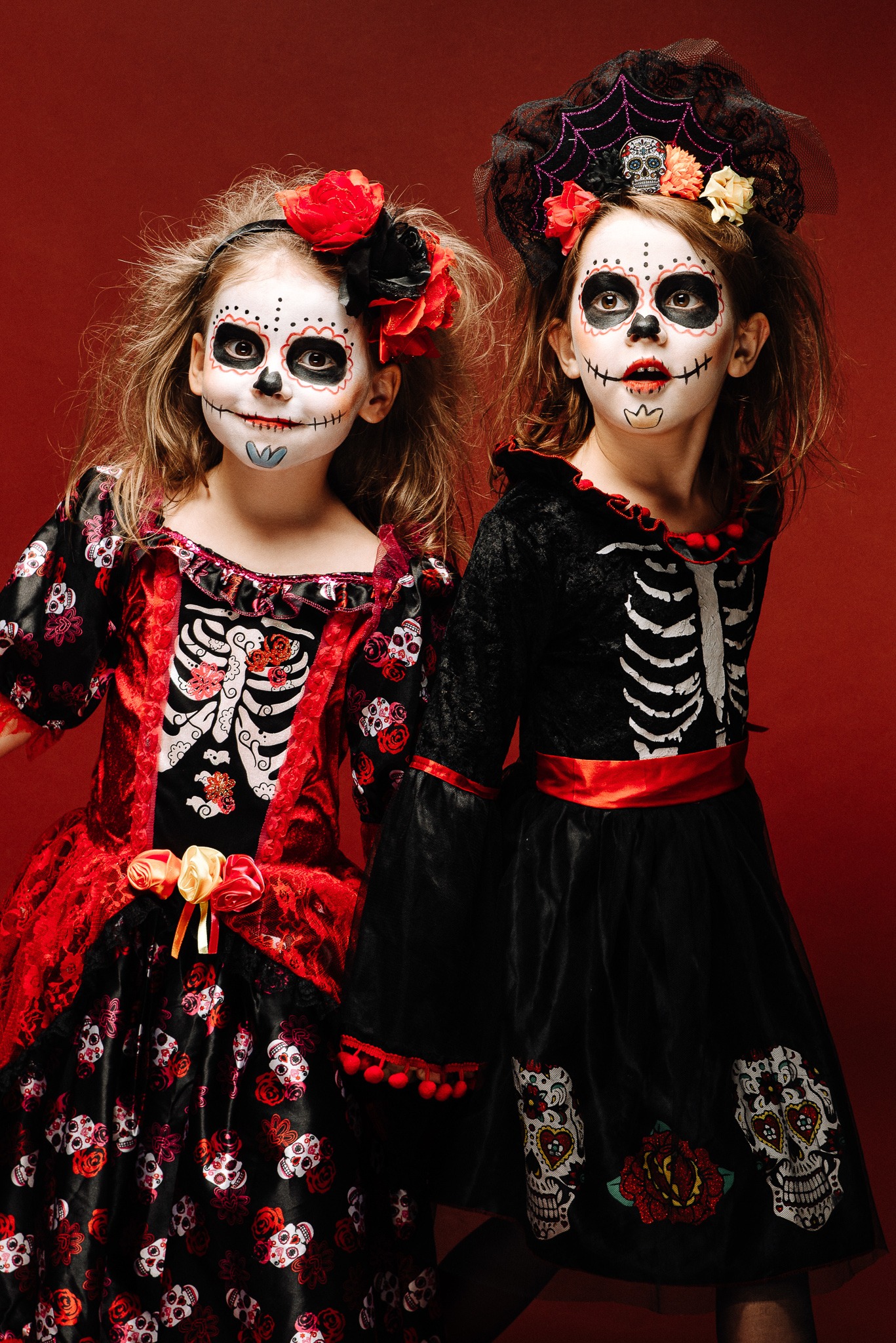 Dwie dziewczynki w kostiumach i makijażu inspirowanym Dniem Zmarłych, na czerwonym tle. Kostiumy zdobione czaszkami i kwiatami, makijaż z motywem szkieletu.