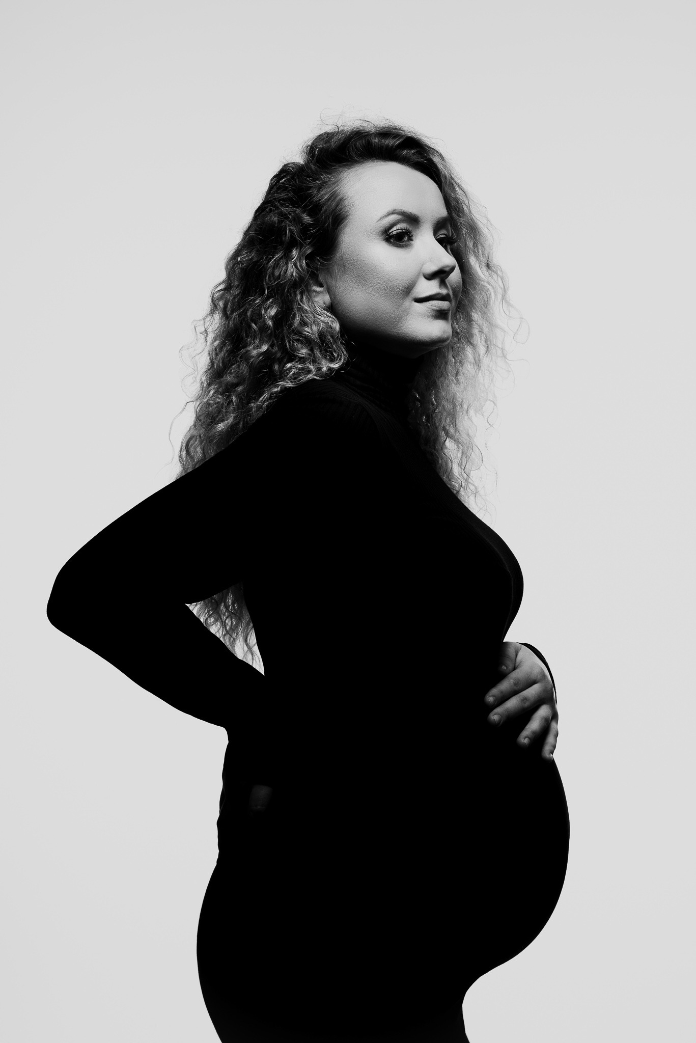 Czarno-białe, studyjne zdjęcie kobiety w ciąży, z kręconymi włosami, ubranej w czarny golf, pozującej z ręką na brzuchu, z profilu, na jasnym tle.