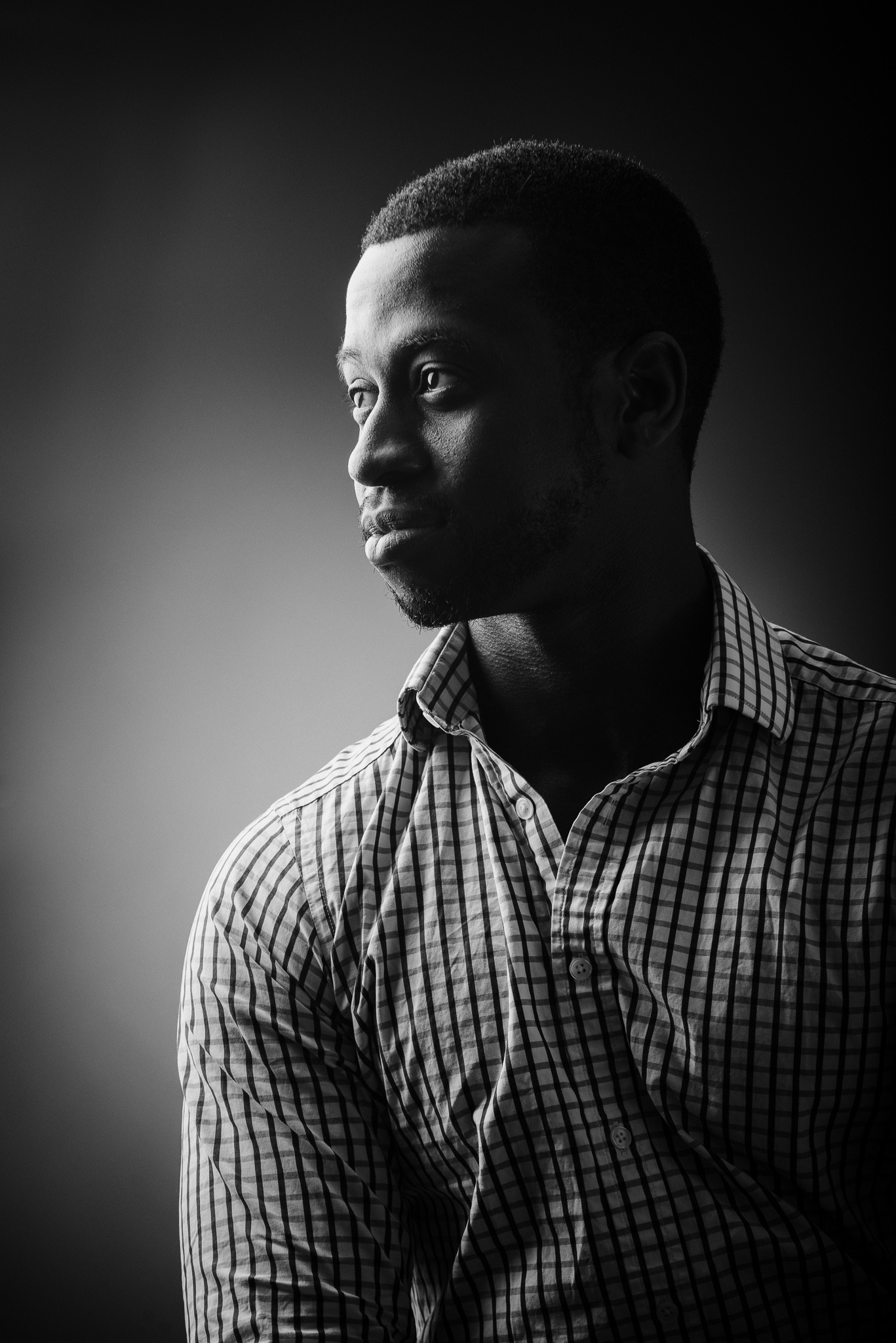 Czarno-białe, profilowe ujęcie mężczyzny w koszuli w kratę, z delikatnym zarostem, spoglądającego w lewo, z miękkim oświetleniem podkreślającym rysy twarzy.
