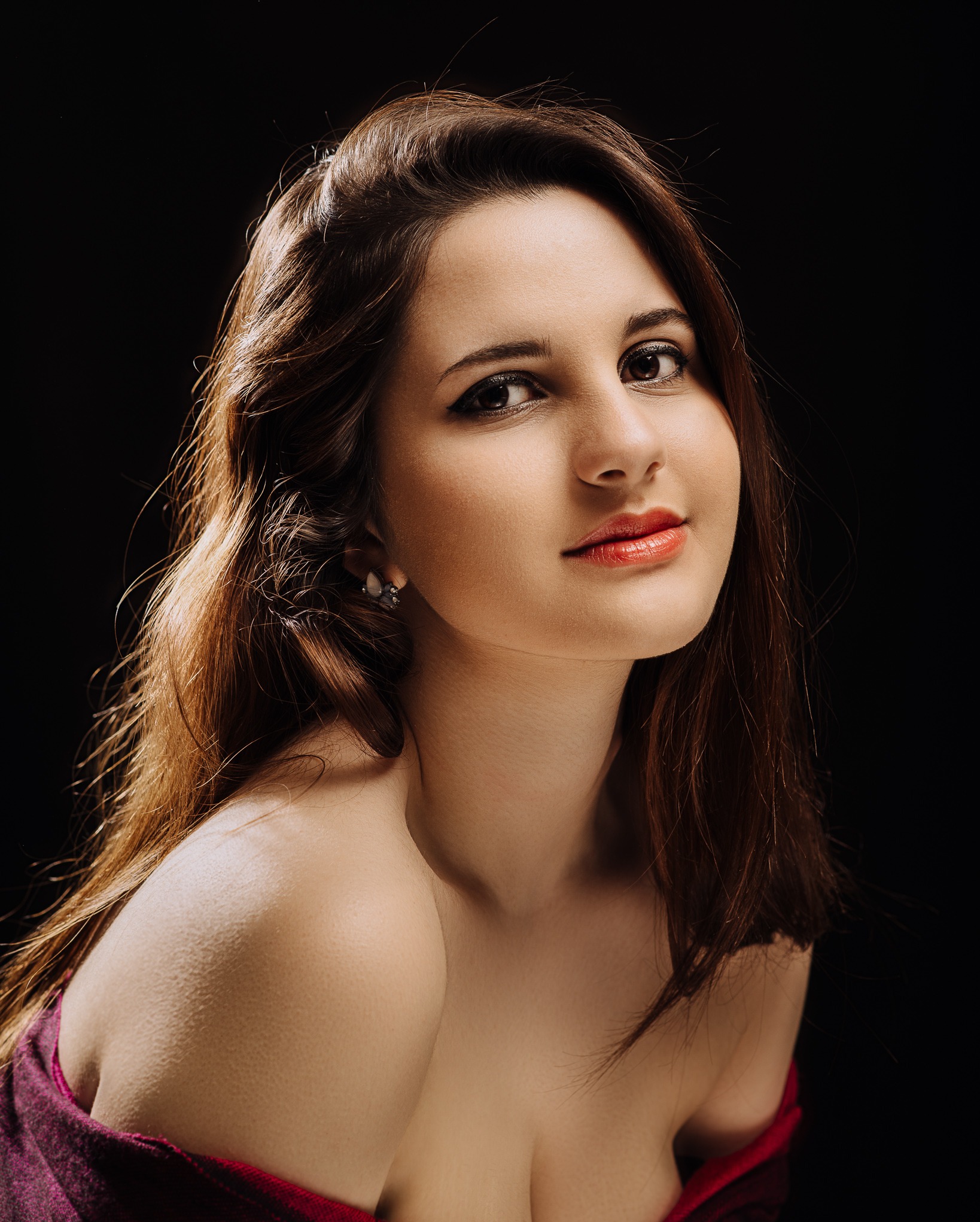 Portret kobiety z długimi, brązowymi włosami, delikatnym makijażem i błyszczącymi ustami na czarnym tle. Widoczny fragment ramienia i biżuteria w uchu.