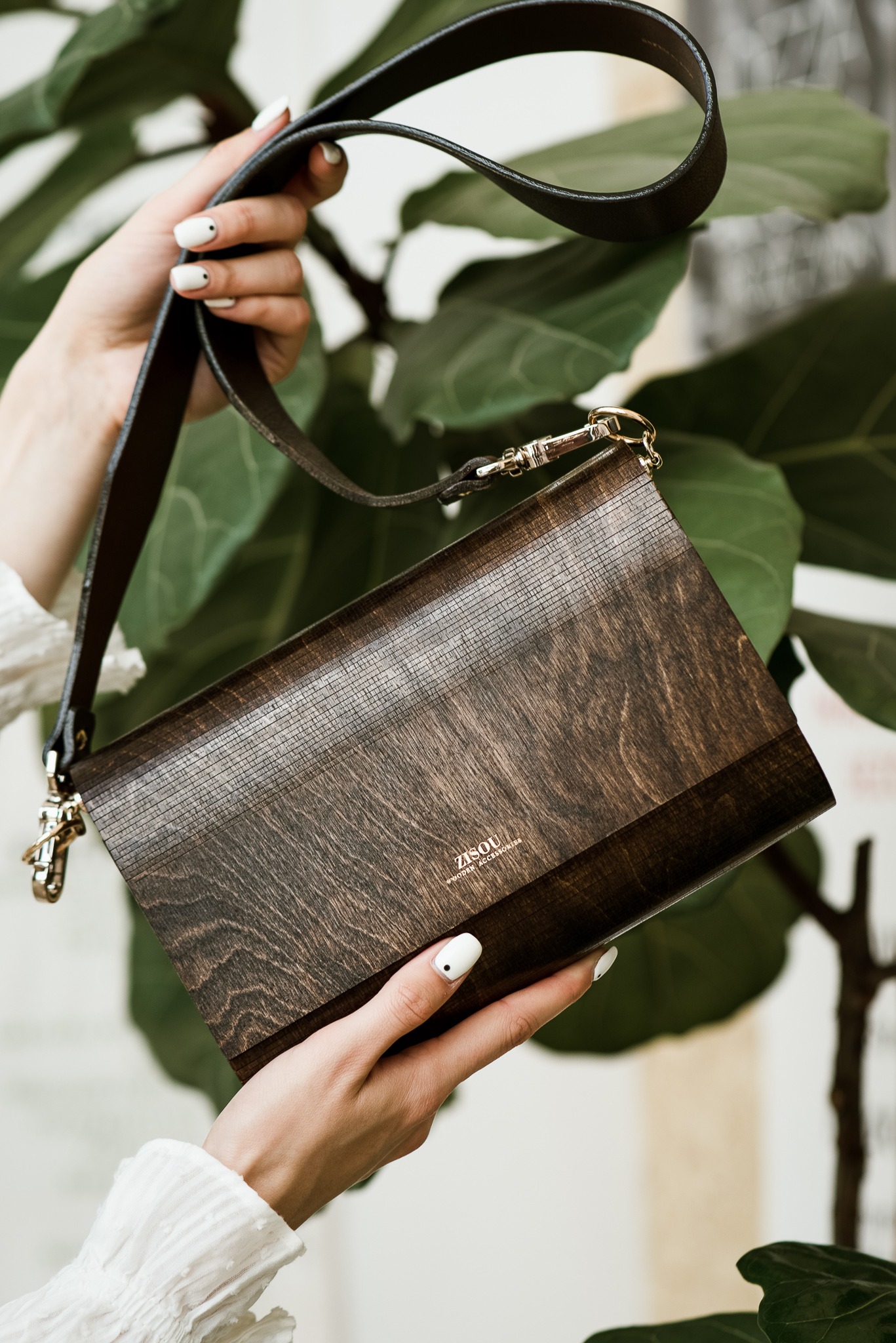 Kobieta prezentuje drewnianą torebkę marki Zisou na tle zielonych liści. Widoczne dłonie z białym manicure i fragment białej bluzki. Elegancki dodatek.