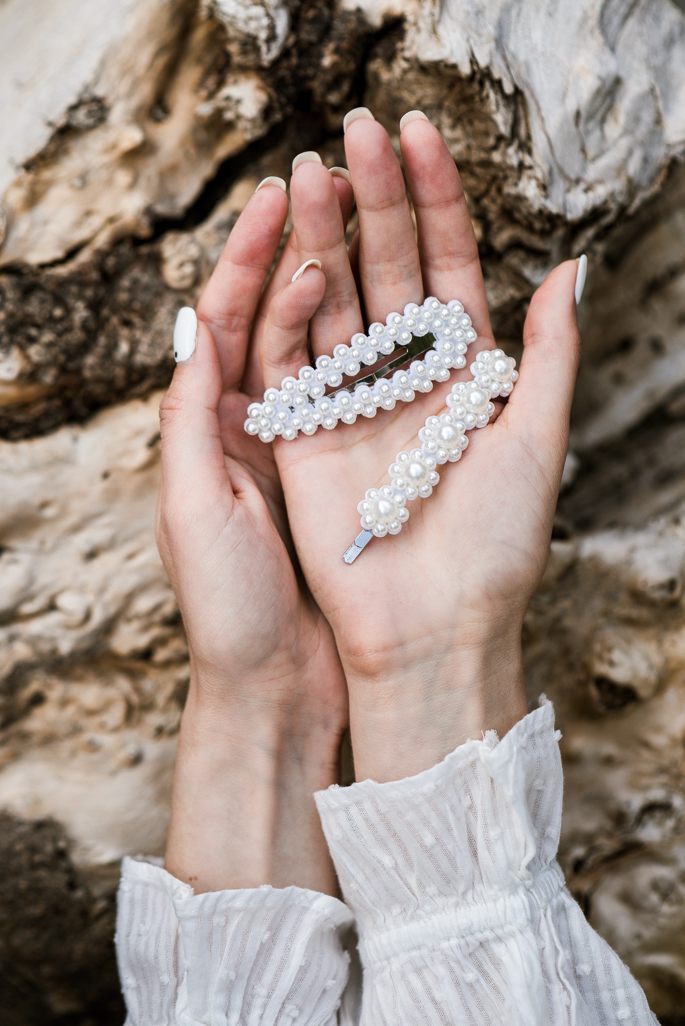 Dwie spinki do włosów z perłami w dłoniach kobiety na tle kory drzewa. Biały manicure i bluzka z falbankami dodają elegancji. Delikatne światło podkreśla detale.