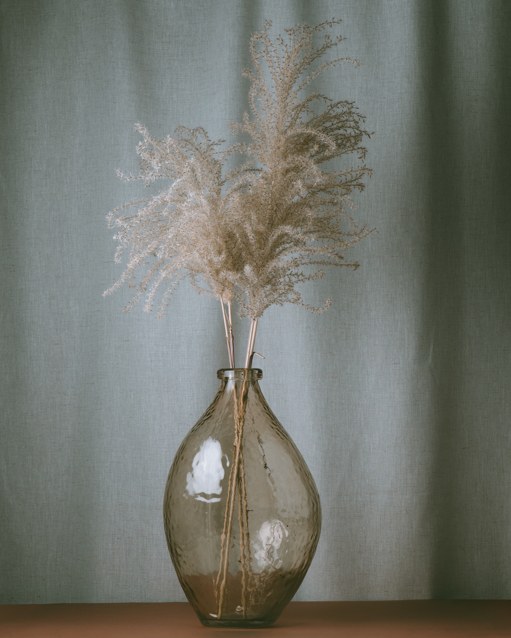 Dekoracyjna, pękata, szklana butelka z suchymi trawami na tle tekstylnej ściany. Stonowana kolorystyka, miękkie światło, minimalistyczny styl.