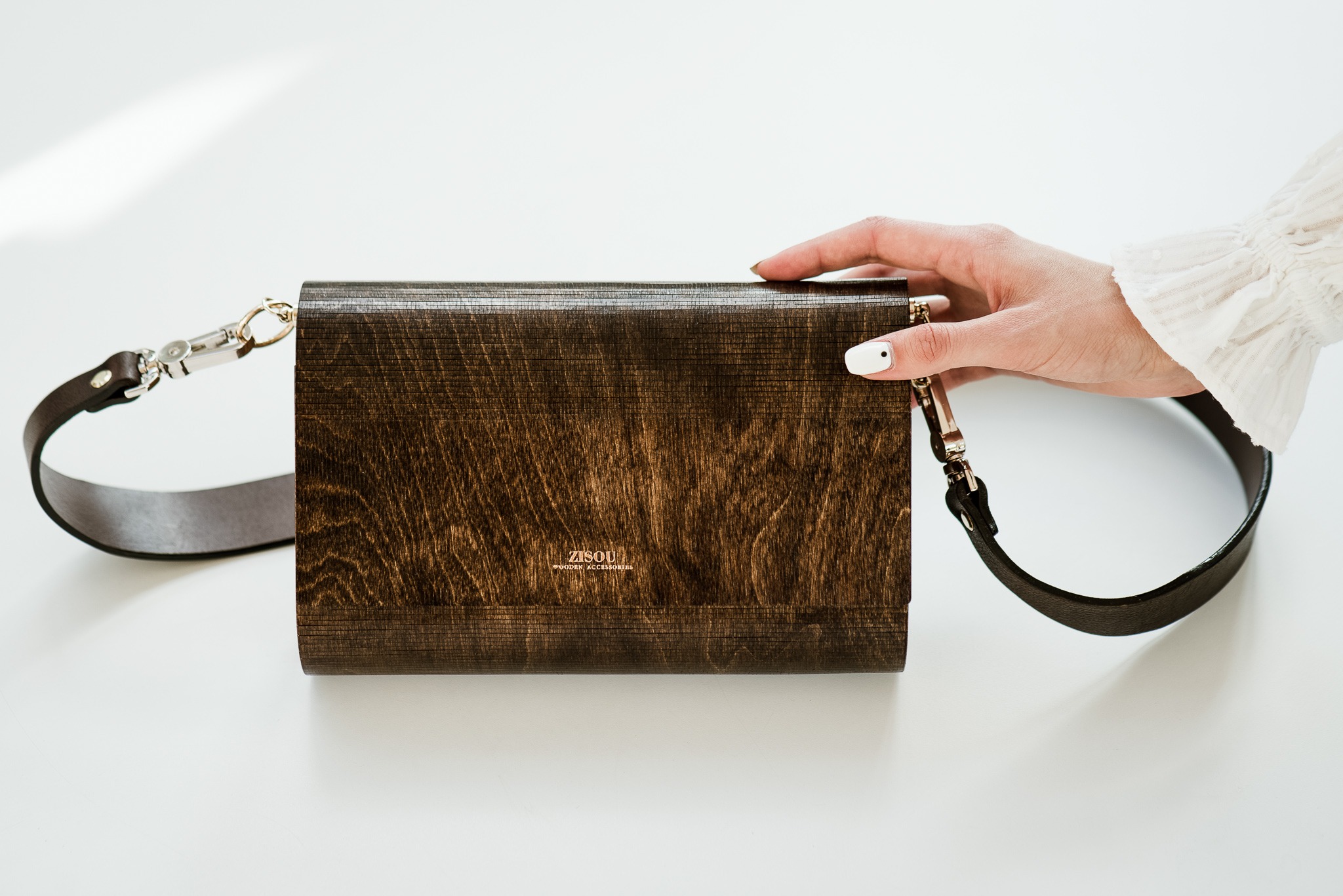 Zdjęcie z góry: dłoń w mankiecie bluzki dotyka drewnianej torebki Zisou z brązowym paskiem na białym tle. Minimalizm, naturalne światło, fotografia produktowa.