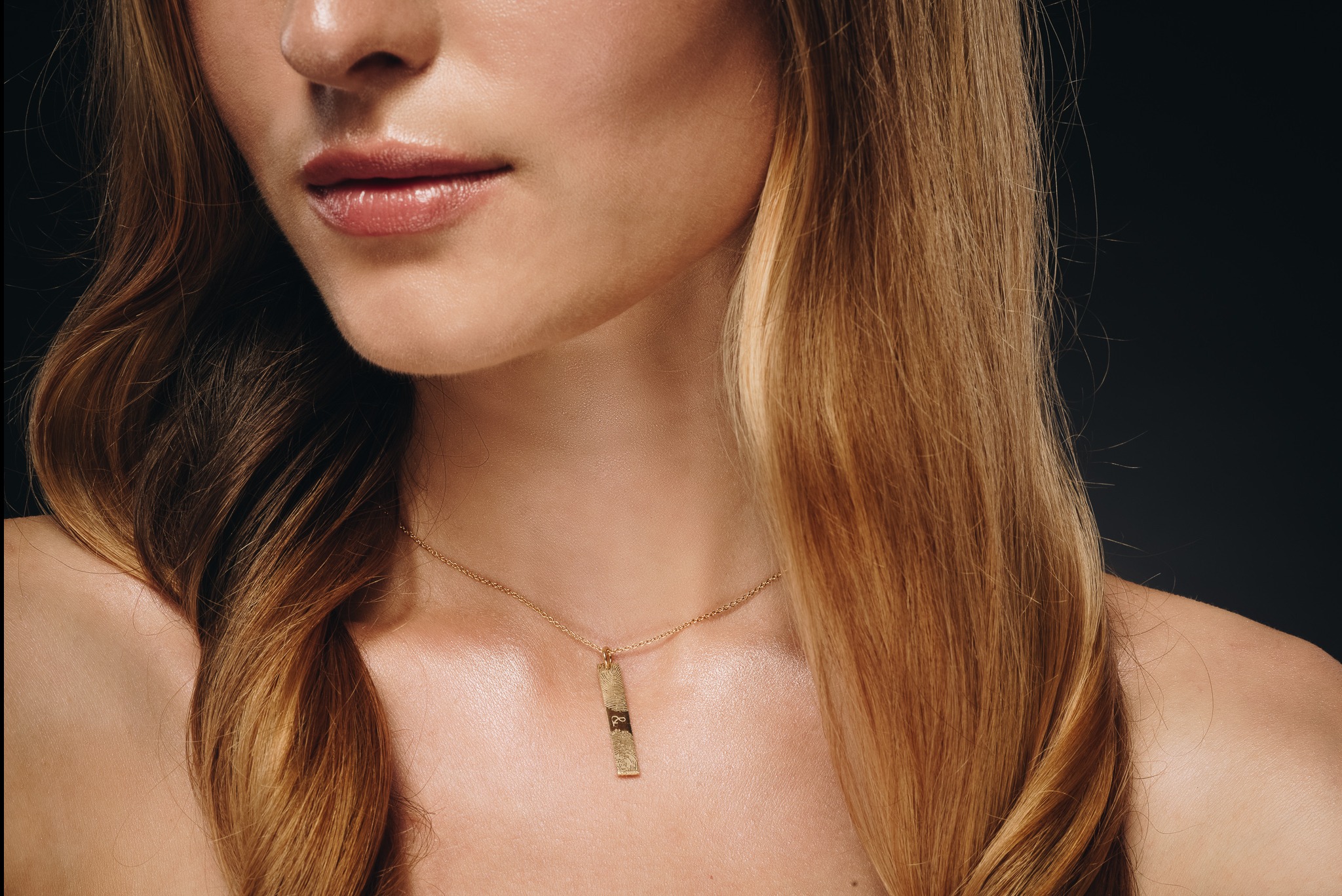 Zbliżenie na szyję modelki z delikatnym złotym łańcuszkiem z zawieszką w kształcie prostokąta z odciskiem palca na ciemnym tle. Subtelny makijaż i blond włosy.
