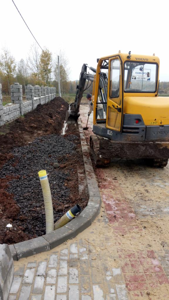 Żółta koparka Volvo EC25 w trakcie wykonywania wykopu pod rury kanalizacyjne, widoczne ułożone obrzeża chodnikowe i fragment muru z bloczków betonowych w tle.