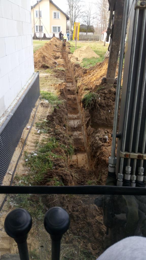 Widok z kabiny koparki na wykopany wąski rów w ziemi, prawdopodobnie pod fundament budynku, z widocznymi śladami opon koparki i fragmentem izolacji fundamentu przy ścianie budynku.