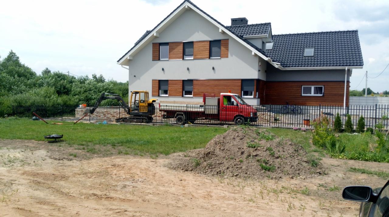 Żółta mini koparka i czerwony wywrotka na tle nowoczesnego domu z drewnianymi elementami, widoczny wykop i sterta ziemi, sugerujące prace ziemne.