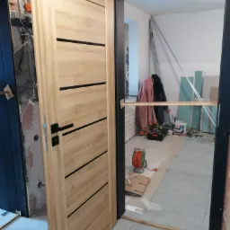 Montaż drzwi wewnętrznych: widok na otwarte, drewniane drzwi z czarną klamką w surowym wnętrzu, z widocznymi elementami instalacji i narzędziami w tle.