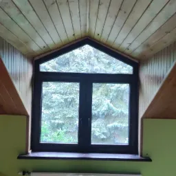 Okno PCV z czarną ramą, widok na zaśnieżone drzewa, drewniana boazeria na suficie i ścianach pod skosem, zielona ściana i biały grzejnik.
