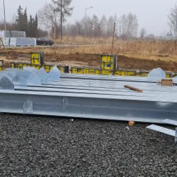 Przygotowane do montażu, ocynkowane elementy konstrukcji stalowej hali na placu budowy, w tle widoczne szalunki fundamentowe i zapakowane płyty warstwowe.