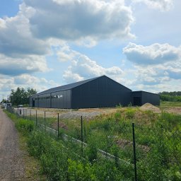 SKELETON SPÓŁKA Z OGRANICZONĄ ODPOWIEDZIALNOŚCIĄ - Gotowa hala magazynowa z ciemnoszarej blachy trapezowej, widok z zewnątrz, ogrodzenie z siatki, częściowo zagospodarowany teren.