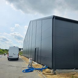 SKELETON SPÓŁKA Z OGRANICZONĄ ODPOWIEDZIALNOŚCIĄ - Gotowa hala z szarej blachy trapezowej, widoczny fragment elewacji z drzwiami, biały bus w tle, niebieskie węże na ziemi.