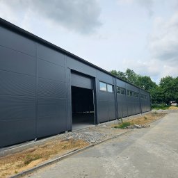 SKELETON SPÓŁKA Z OGRANICZONĄ ODPOWIEDZIALNOŚCIĄ - Szara hala z otwartą bramą segmentową, widoczne okna i teren wokół w trakcie prac wykończeniowych.