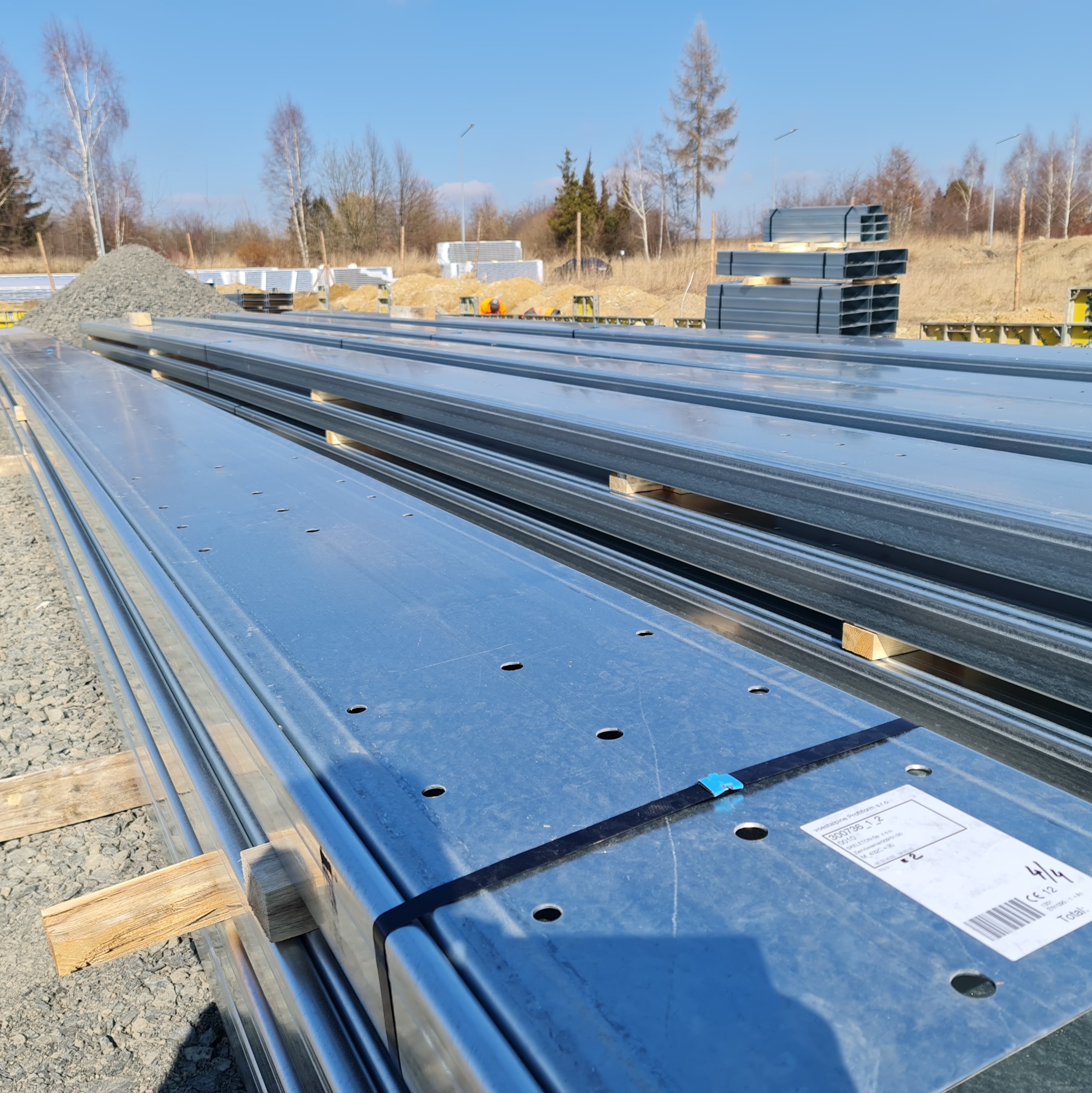 Stos ocynkowanych profili stalowych z otworami, zabezpieczonych czarną taśmą i etykietą producenta, składowanych na placu budowy na drewnianych legarach.