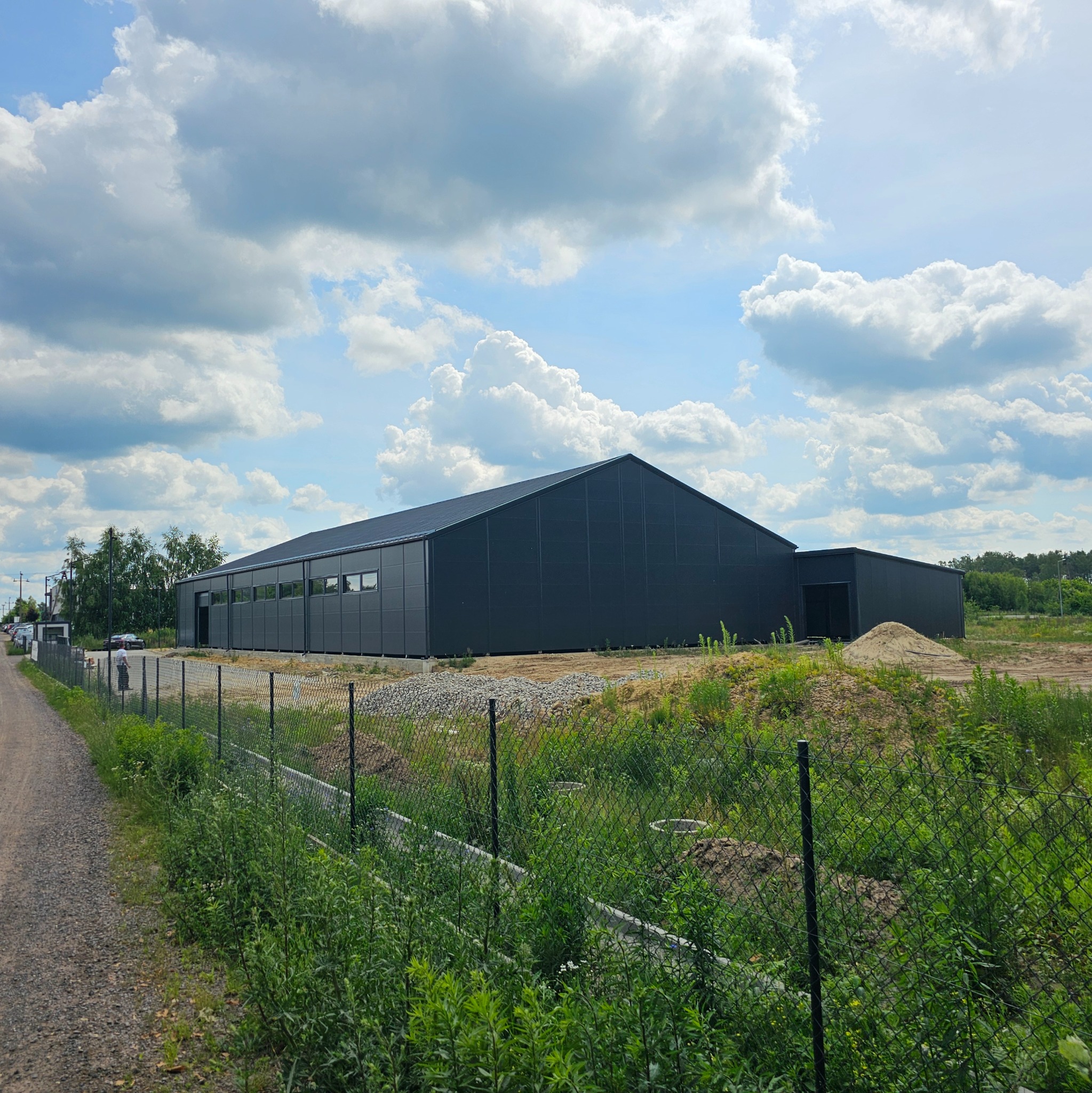 Gotowa hala magazynowa z ciemnoszarej blachy trapezowej, widok z zewnątrz, ogrodzenie z siatki, częściowo zagospodarowany teren.