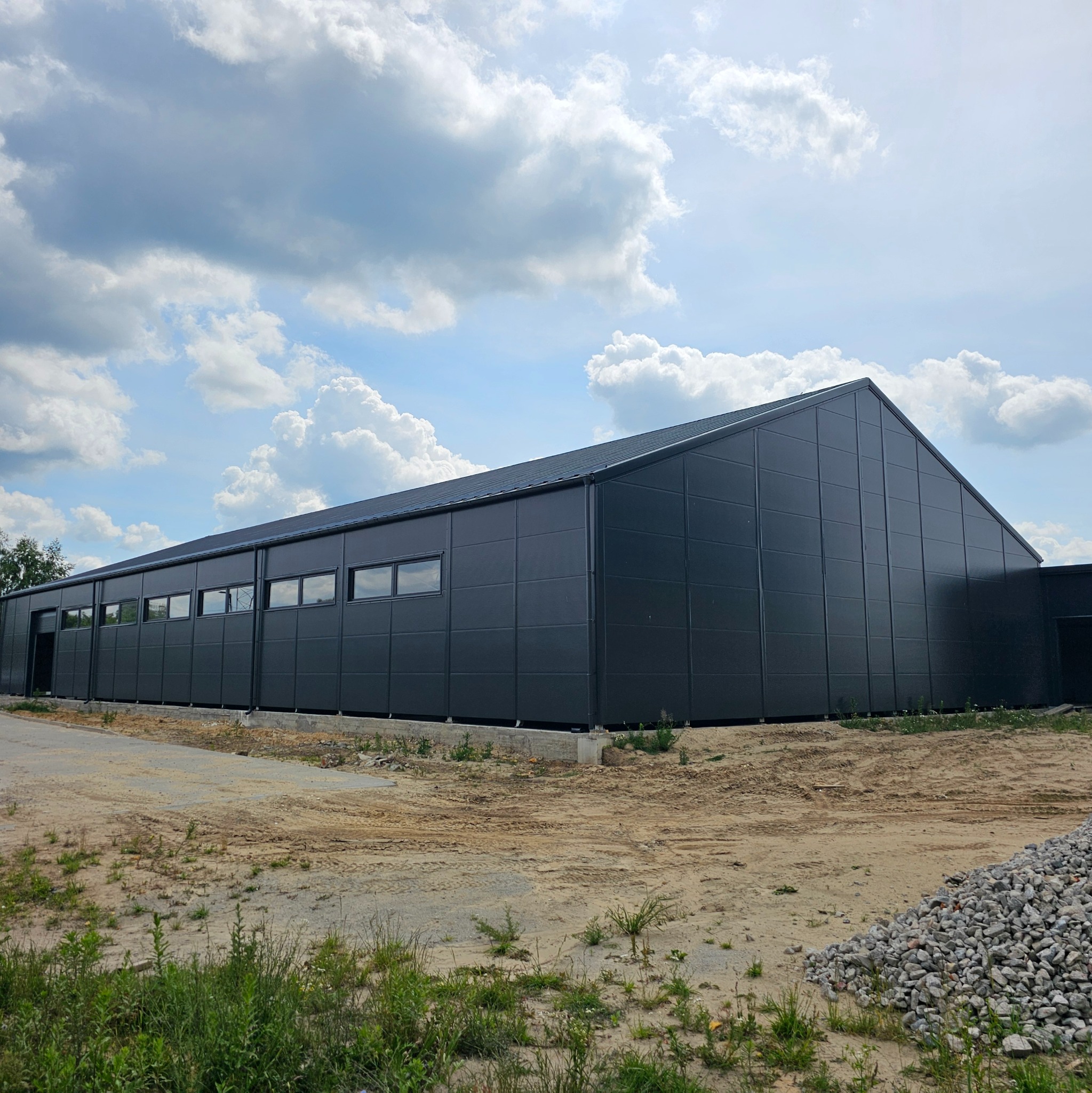 Gotowa hala stalowa w kolorze grafitowym, widok z zewnątrz, z widocznymi oknami i bramą wjazdową, na tle błękitnego nieba z obłokami.