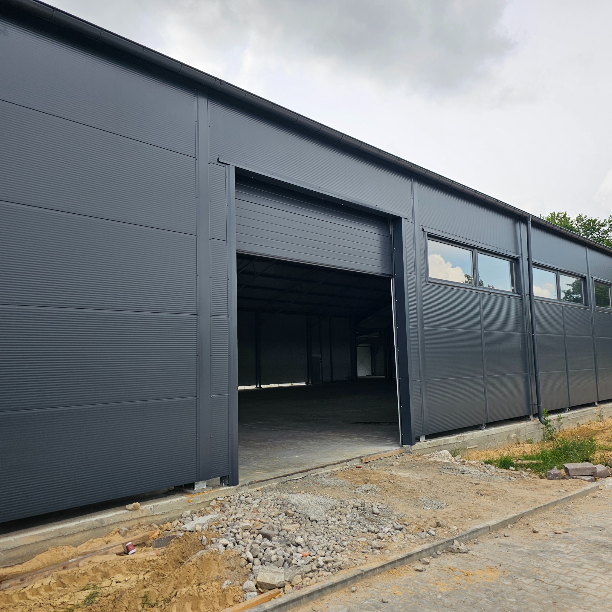 Szara hala z blachy falistej z otwartą bramą rolowaną i widocznym wnętrzem, teren wokół w trakcie zagospodarowania.