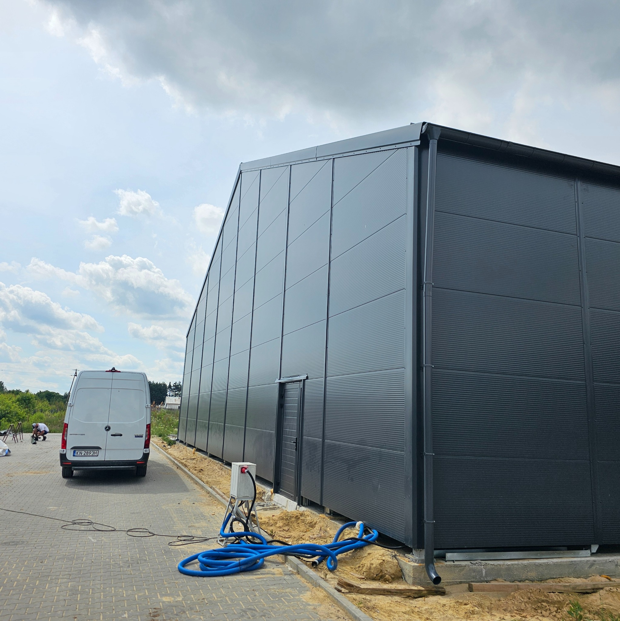 Gotowa hala z szarej blachy trapezowej, widoczny fragment elewacji z drzwiami, biały bus w tle, niebieskie węże na ziemi.