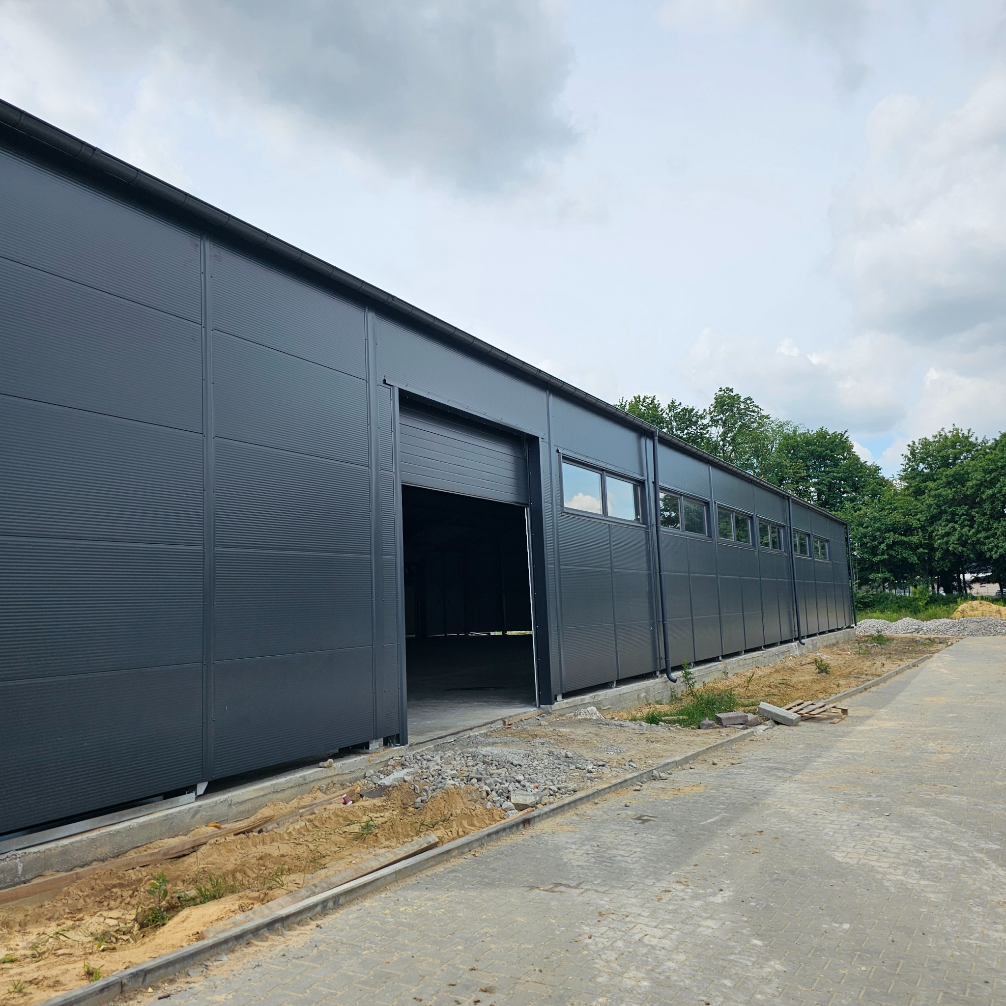 Szara hala z otwartą bramą segmentową, widoczne okna i teren wokół w trakcie prac wykończeniowych.