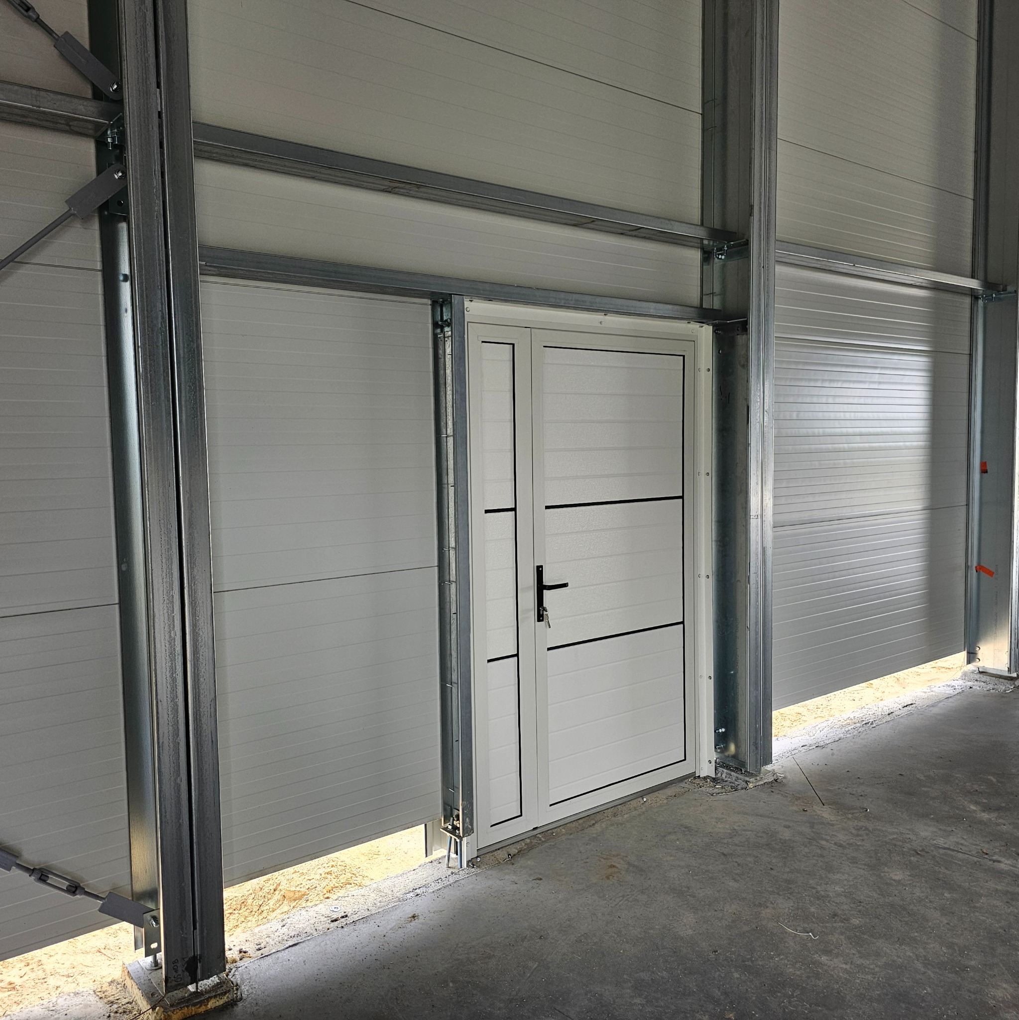 Wnętrze hali w trakcie budowy, widoczne białe panele ścienne, stalowa konstrukcja nośna oraz dwuskrzydłowe białe drzwi z czarnymi akcentami i klamką, z kluczem w zamku.