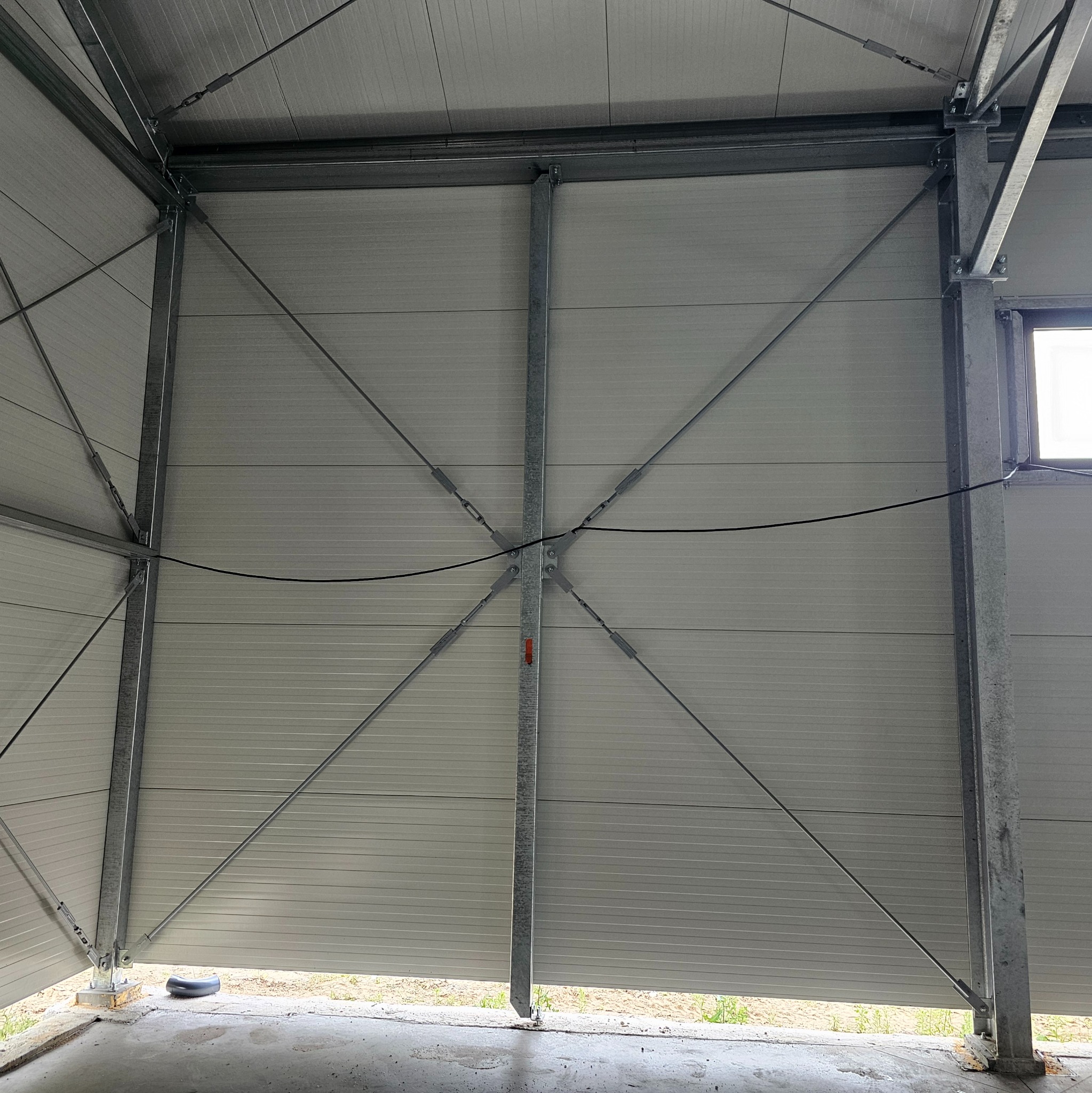 Wnętrze hali w trakcie budowy, widoczne stalowe elementy konstrukcyjne i panele ścienne, mocowania śrubowe, zarys okna w tle.