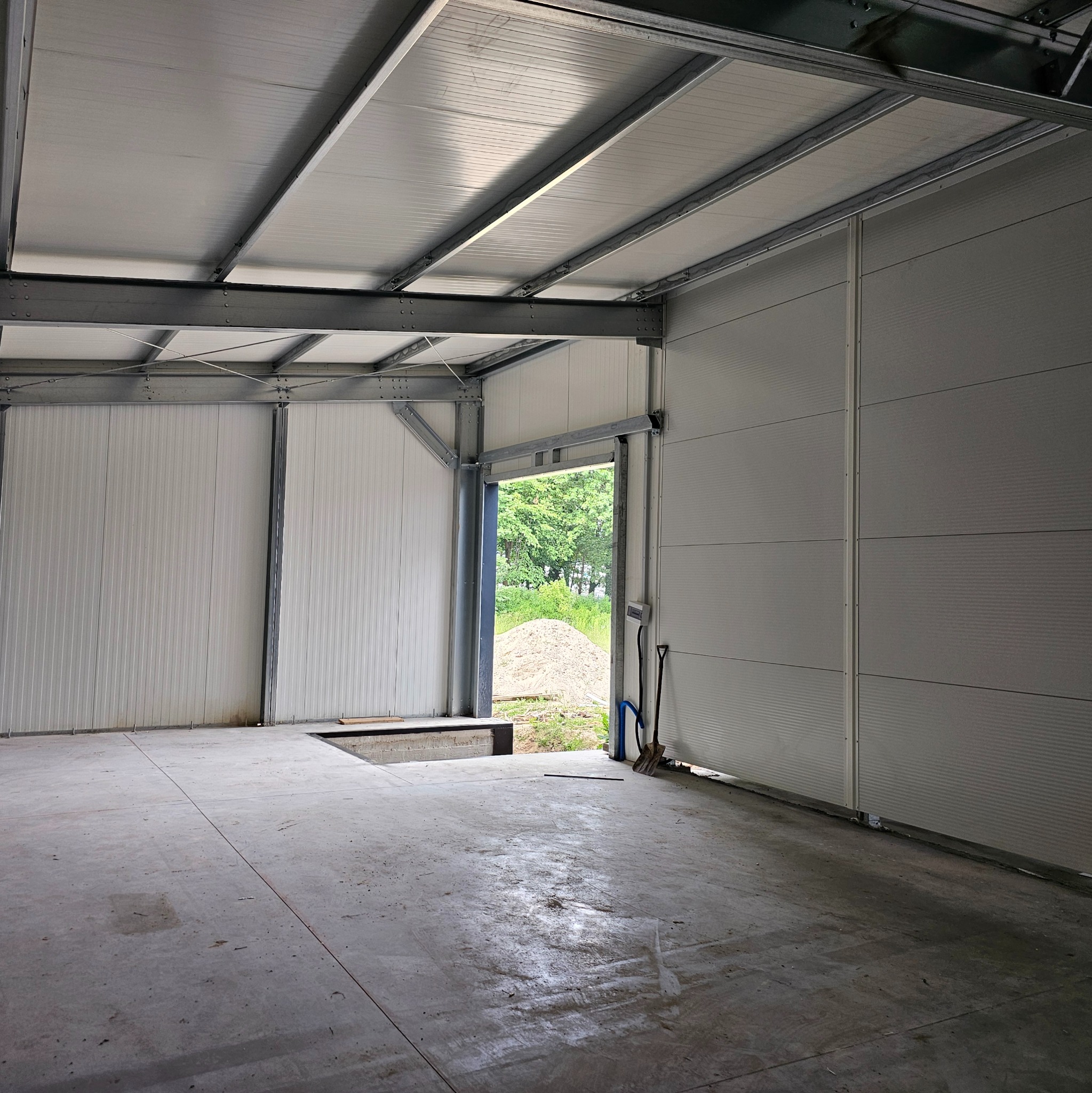 Wnętrze nowo wybudowanej hali z widoczną stalową konstrukcją, ścianami z paneli warstwowych i betonową posadzką; przez otwarty wjazd widać hałdę piasku i zieleń.