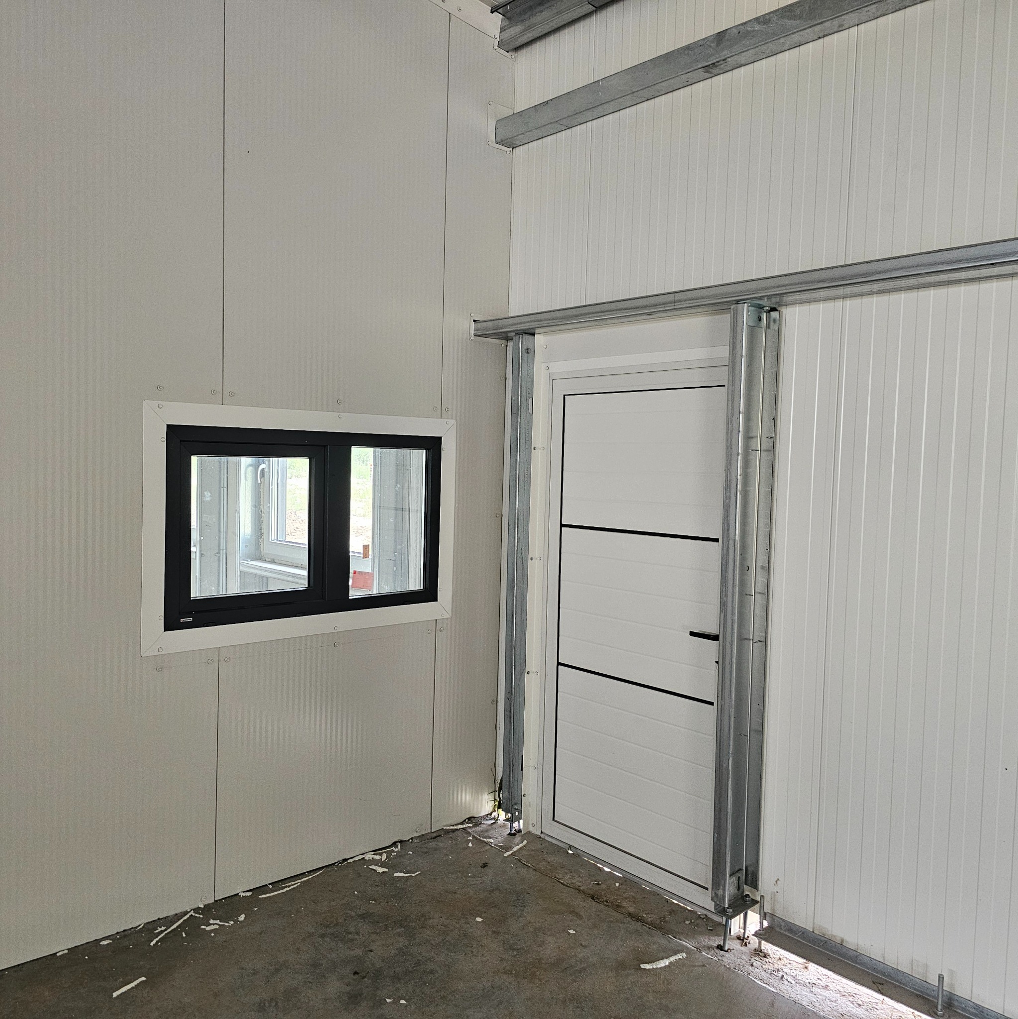 Wnętrze hali w trakcie budowy z widocznym oknem w czarnej ramie i przesuwnymi drzwiami garażowymi na metalowej szynie, surowy betonowy podkład z resztkami pianki montażowej.