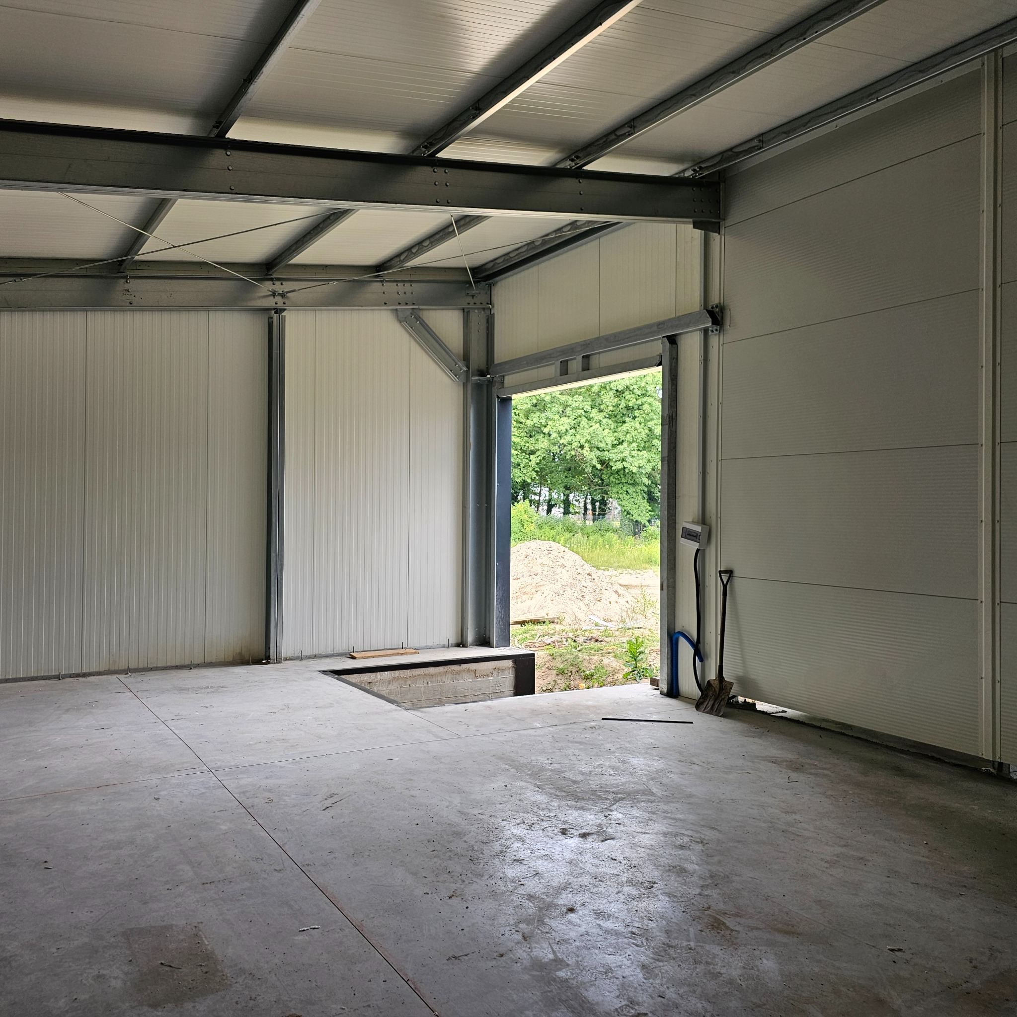 Wnętrze hali w trakcie budowy, widoczne stalowe elementy konstrukcyjne, ściany z paneli, betonowa posadzka z wykopem, otwarte wrota z widokiem na teren zewnętrzny i hałdę piachu, łopata oparta...