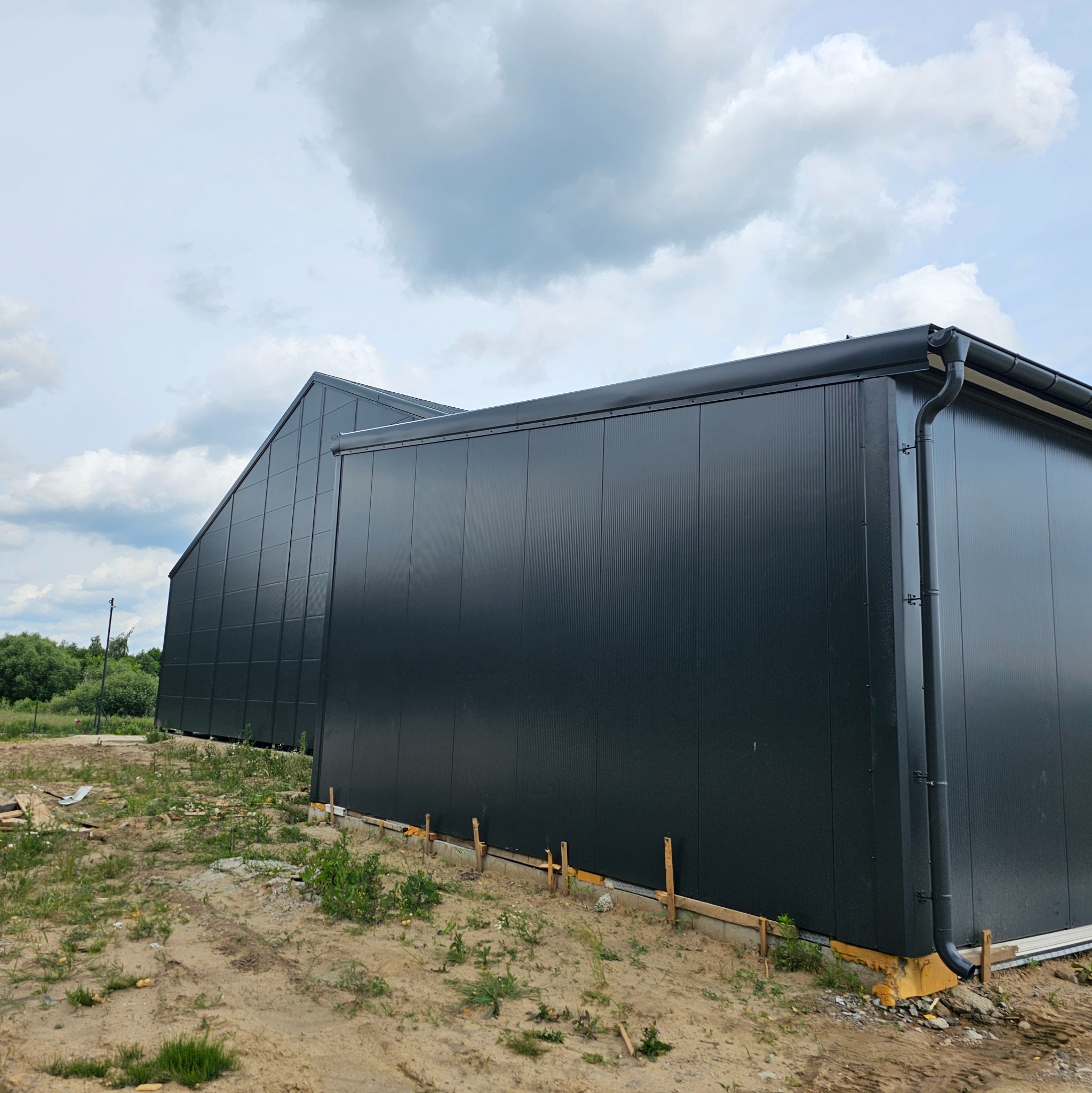 Dwie połączone hale w kolorze antracytowym, wykonane z paneli warstwowych, widoczne na etapie wykończenia terenu wokół budynków. Widoczne drewniane szalunki fundamentów.