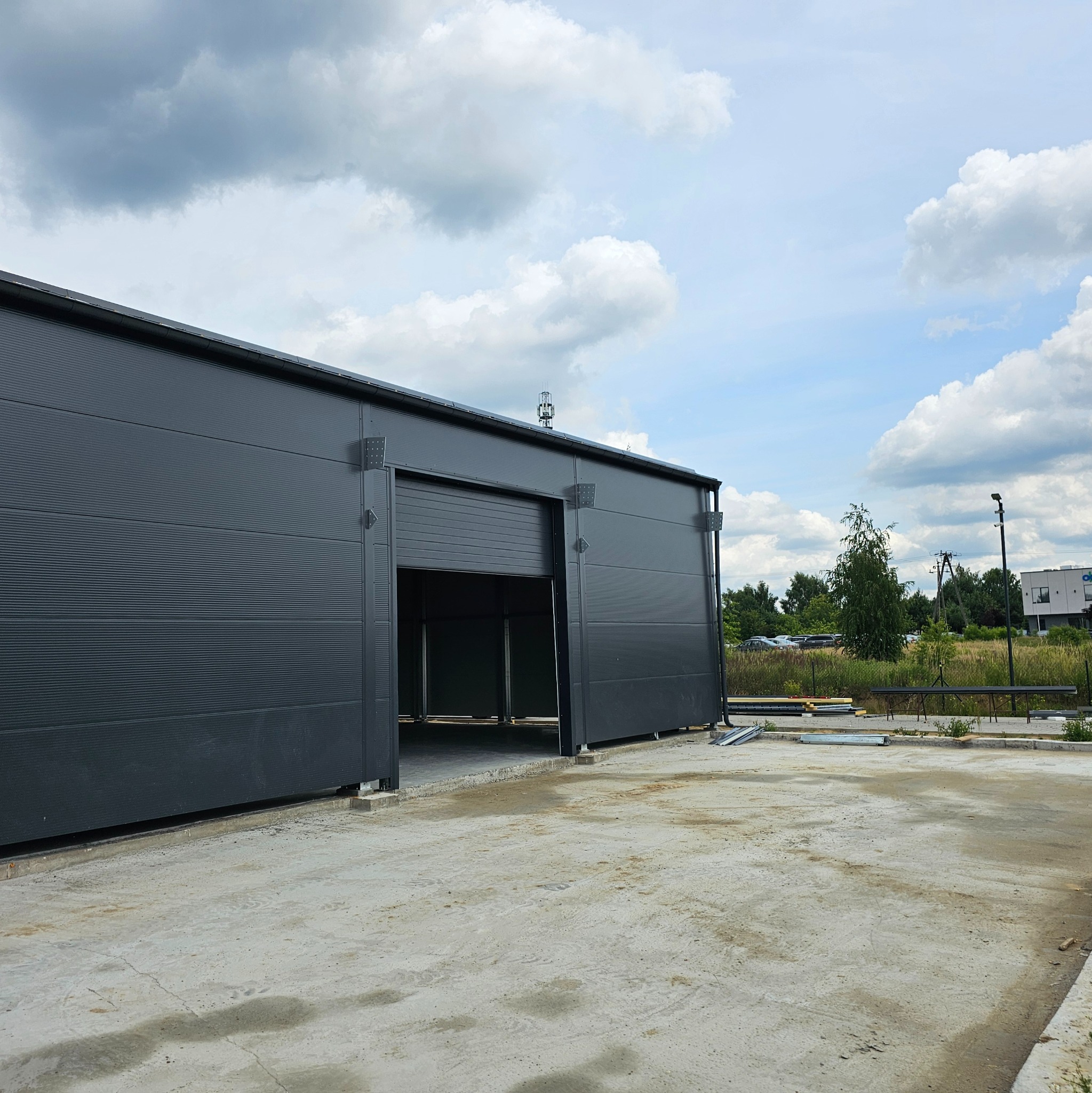 Częściowo otwarta brama wjazdowa do szarej hali stalowej z widoczną konstrukcją mocującą, na betonowym placu, w tle zieleń i pochmurne niebo.