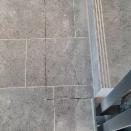 Ułożona szara, matowa terakota na podłodze i schodach przy metalowej barierce, widoczny fragment buta roboczego.