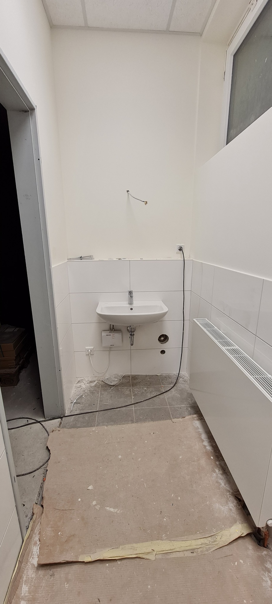 Łazienka w trakcie remontu: białe płytki ścienne, umywalka zamontowana, widoczne przewody, grzejnik panelowy, podłoga przygotowana pod wylewkę.
