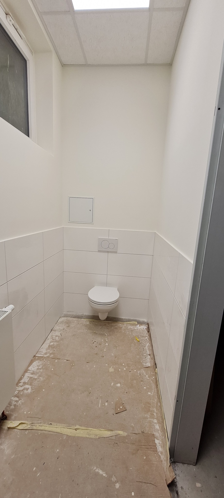 Łazienka w trakcie remontu: białe płytki ścienne, wisząca toaleta, surowa podłoga przygotowana pod wylewkę, widoczna klapka rewizyjna.
