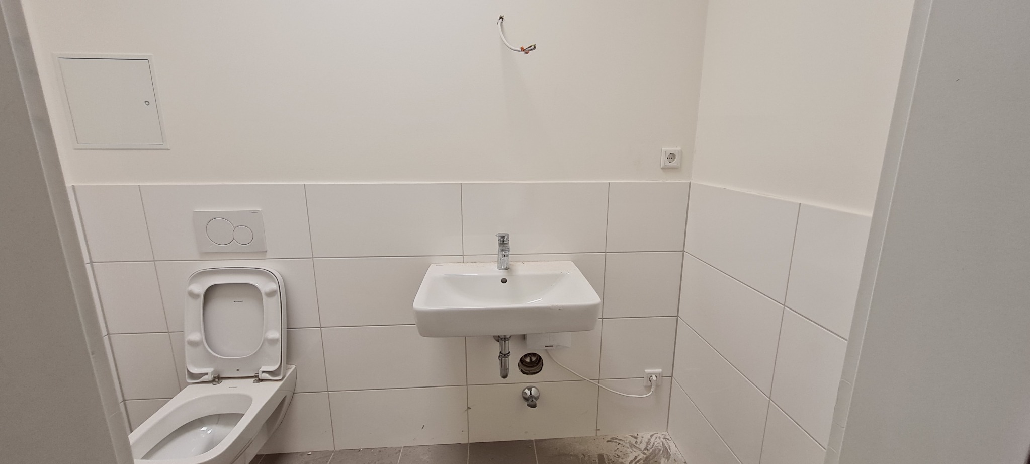 Wnętrze łazienki w trakcie prac wykończeniowych, widoczna umywalka z baterią, toaleta z otwartą klapą oraz białe płytki na ścianach. Na ścianie widoczne wystające kable instalacji elektrycznej.